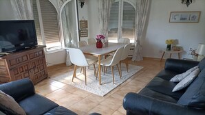 A living room with a TV, sofa, dining table, chairs, and a small side table with a lamp.