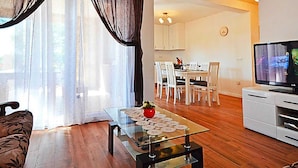 Un salon moderne avec une table basse en verre, un plancher en bois, un canapé, un coin repas avec des chaises, un téléviseur et une cuisine en arrière-plan.