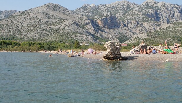 Une plage où l’on voit des gens nager et se détendre, une structure en pierre et, au loin, des montagnes.