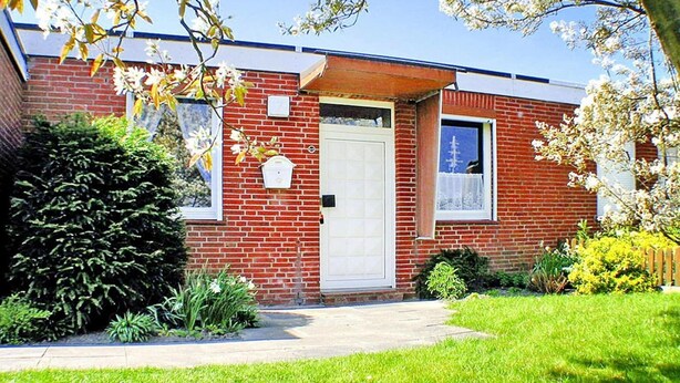 A brick house with a white door, a small porch, and a garden with various plants. - Gemütlicher Bungalow Kabeljau in Dornumersiel (Dornumersiel)