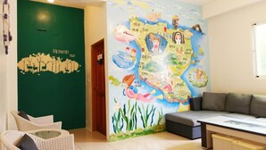 Une pièce avec un mur vert, une fresque murale sur le thème des sirènes et du monde sous-marin, un canapé gris et des fauteuils en osier.