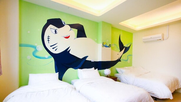 Une chambre d’hôtel avec trois lits, une grande fresque murale représentant un personnage de dessin animé et un climatiseur fixé au mur.