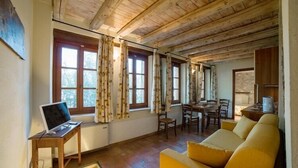 Une pièce avec des poutres apparentes en bois au plafond, un canapé jaune, une table à manger avec des chaises et un ordinateur portable posé sur une petite table.