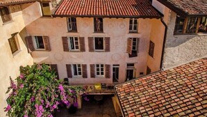 Un immeuble à plusieurs étages, doté d’un toit recouvert de tuiles rouges, de nombreuses fenêtres avec volets et d’une cour intérieure où pousse une plante à fleurs.