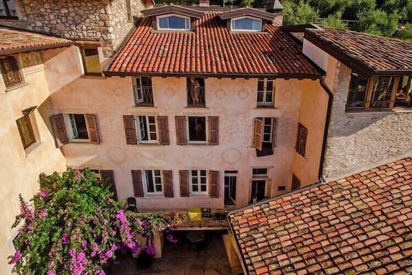 Um edifício de vários andares com telhado de telhas vermelhas, várias janelas com persianas e um pátio com uma planta florida.
