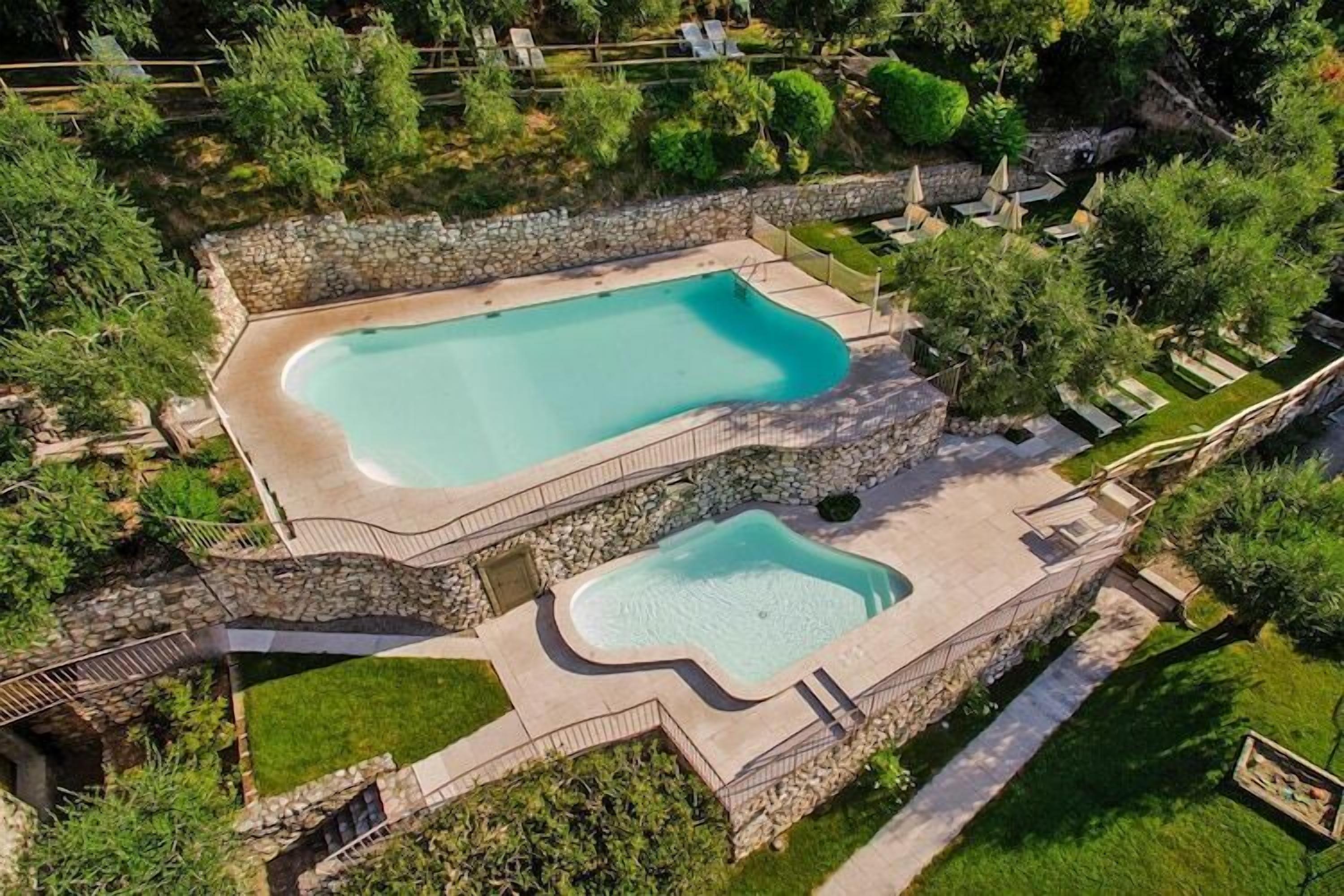 A large rectangular swimming pool with a stone border, surrounded by a stone wall and lush greenery.