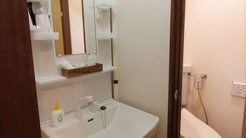 A bathroom with a white sink, mirror, and toilet. There are shelves above the sink and a pair of slippers on the floor.