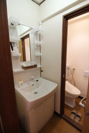 A bathroom with a white sink, mirror, and toilet. There are shelves above the sink and a pair of slippers on the floor. - Guesthouse BJ STYLES (Beppu)