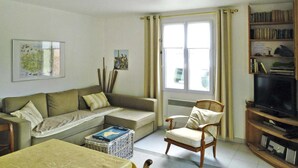 A living room with a sofa, armchair, television, bookshelf, and a window with curtains. - Beachside Flat with Sunny Patio (Cayeux-sur-Mer)