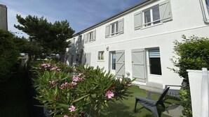 A two-story white house with shutters, a small garden, and a patio chair. - Beachside Flat with Sunny Patio (Cayeux-sur-Mer)