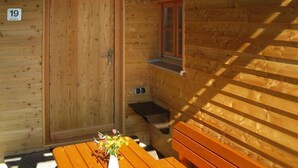 Eine Holzhütte mit einer Tür, auf der '19' steht, einem kleinen Fenster, einer Bank und einem Tisch mit einem Blumenstrauß.