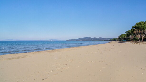 Una amplia playa de arena con aguas tranquilas, cielo despejado y colinas a lo lejos.