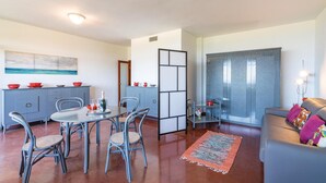 Una sala de estar moderna con un sofá gris, un comedor con una mesa redonda y sillas, una cocina con gabinetes grises y un ventanal con cortinas.