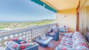 Un balcón con mobiliario de mimbre, una mesa con vasos y botellas, y vistas al mar y las colinas.