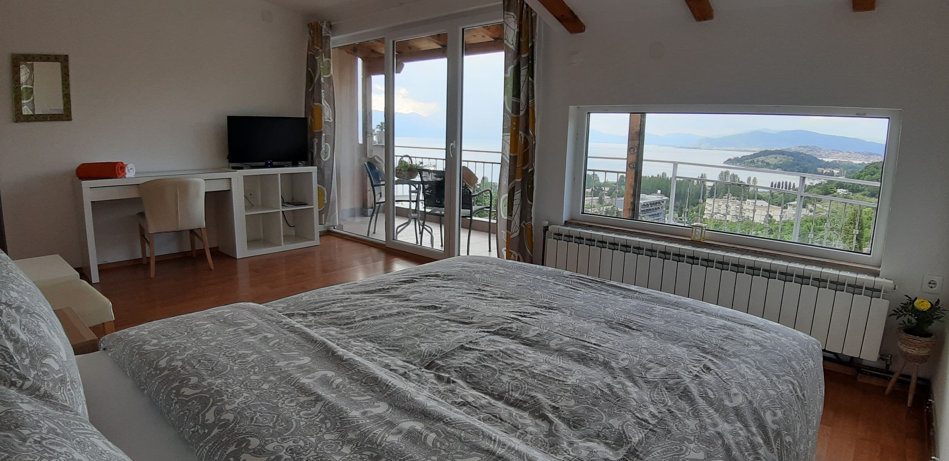 A bedroom with a large bed, a desk with a TV, and a balcony with a view of a lake and mountains.