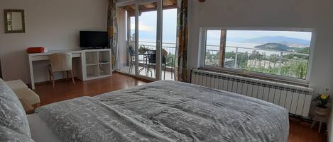 A bedroom with a large bed, a desk with a TV, and a balcony with a view of a lake and mountains.