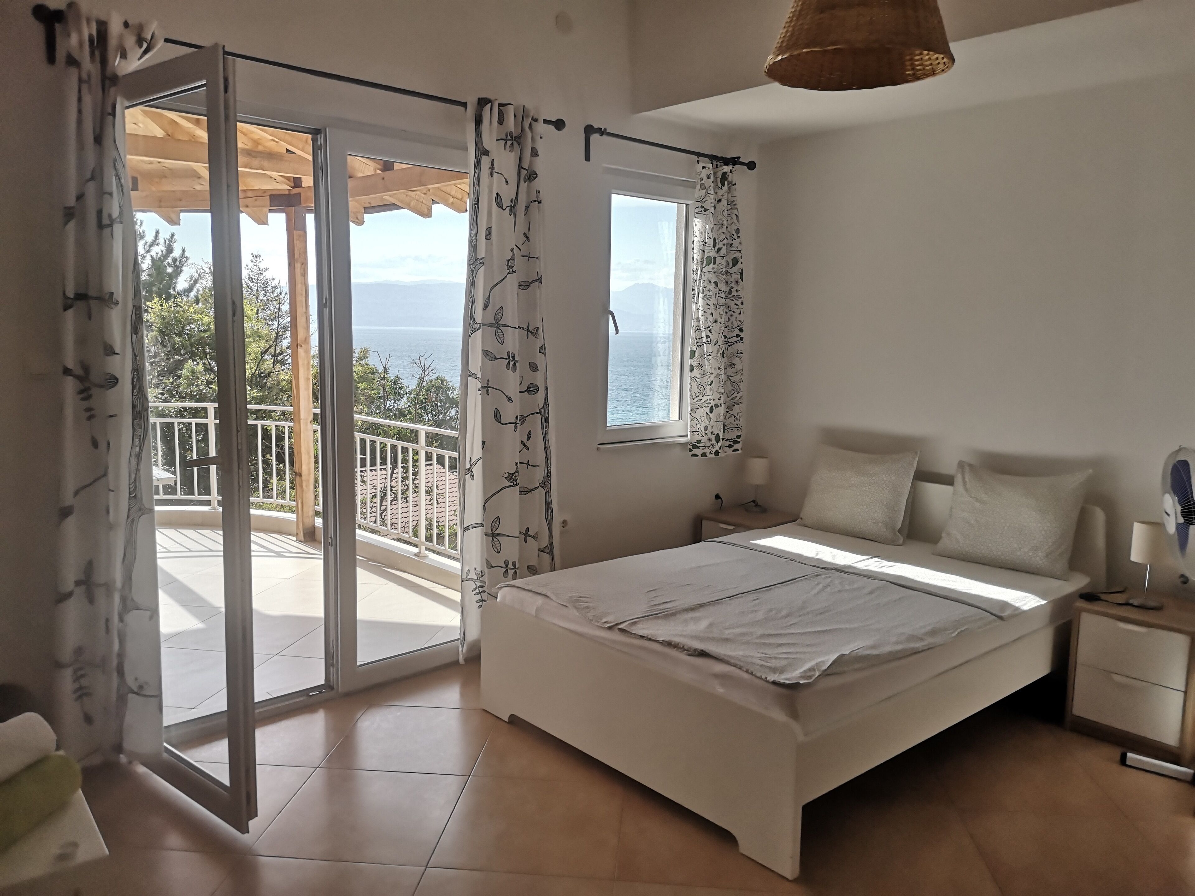 A bedroom with a bed, a nightstand, a lamp, a window with curtains, and a view of the sea.