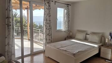 A bedroom with a bed, a nightstand, a lamp, a window with curtains, and a view of the sea.