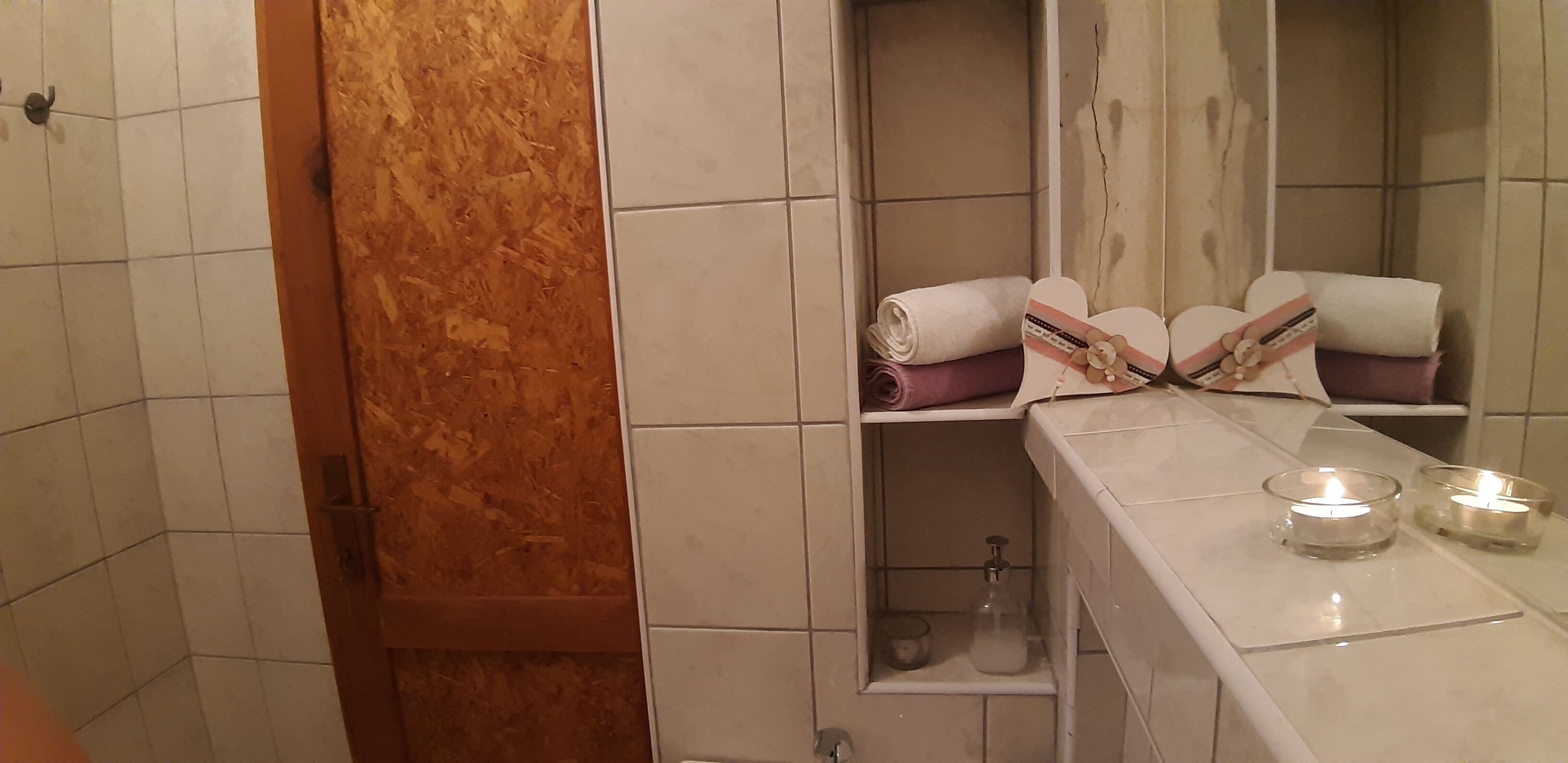 A bathroom with white tiled walls, a wooden door, a mirror, shelves with towels, and two lit candles on the counter.