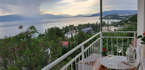 A balcony with a table set for two, overlooking a lake and distant mountains.