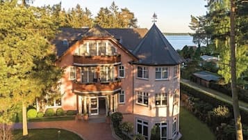 A large, multi-story building with a prominent turret and a balcony, surrounded by lush greenery and a body of water in the background.