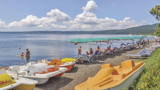 Una spiaggia con file di lettini e ombrelloni, una fila di piccole imbarcazioni e un mare calmo sotto un cielo parzialmente nuvoloso.