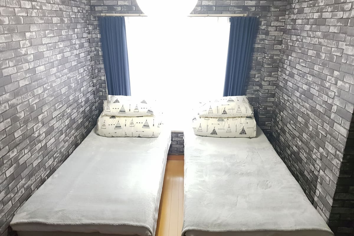 Une chambre avec deux lits, chacun agrémenté d’un ensemble d’oreillers assortis, le tout adossé à des murs en briques, avec une fenêtre au fond.
