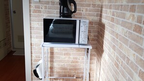 A compact kitchen area with a microwave, kettle, and a small white shelf unit against a brick wall. - Fukuoka Story I (Fukuoka)