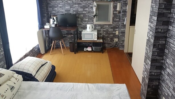 A compact room with a brick-patterned wall, a bed with patterned pillows, a desk with a computer and a kettle, and a microwave. - Fukuoka Story I (Fukuoka)