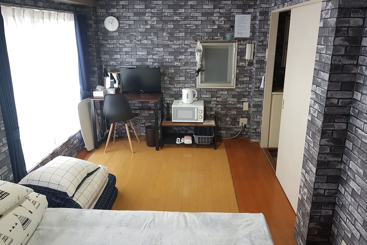 Une pièce de taille modeste avec un mur décoré d’un motif en briques, un lit agrémenté d’oreillers à motifs, un bureau avec un ordinateur et une bouilloire, ainsi qu’un four à micro-ondes.