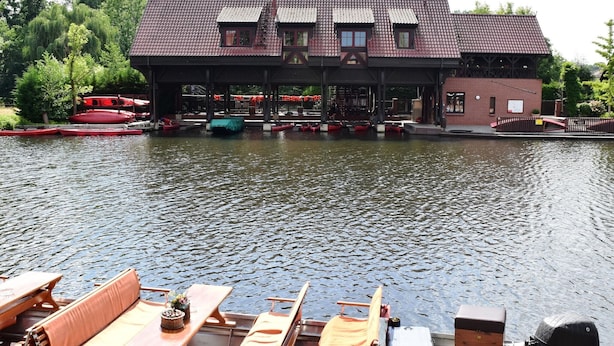 A riverside building with a covered deck area, boats docked in the water, and a row of wooden benches along the riverbank. - Appartements mit Blick auf die Spree in Lübben (Lübben)