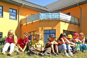 Een groep kinderen zitten op het gras voor een gebouw met een bord waarop wellness en massage diensten worden aangeboden.