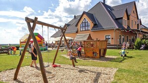 Un parque infantil con columpios y una estructura de juego de madera, frente a una casa con techo de tejas.