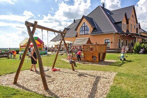 Un parque infantil con columpios y una estructura de juego de madera, frente a una casa con techo de tejas.