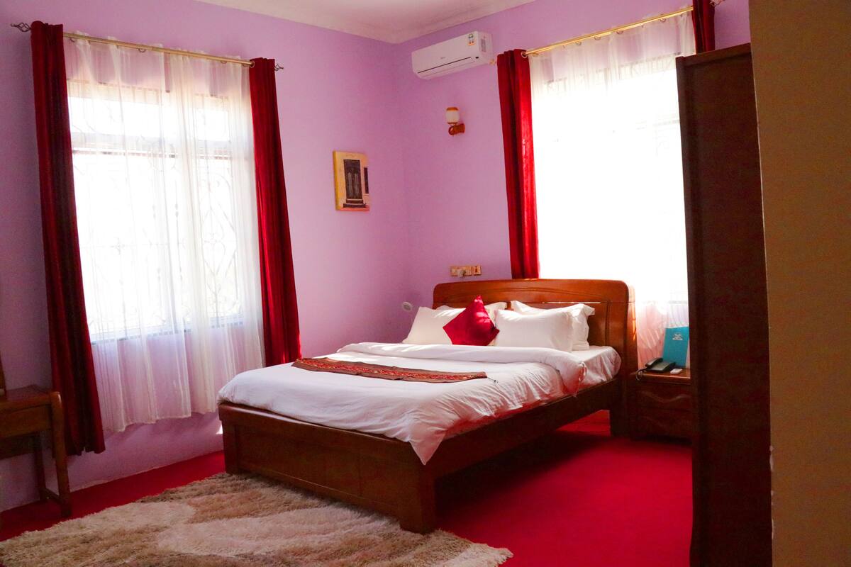 Quarto com cama de madeira, cortinas vermelhas, tapete branco e um aparelho de ar condicionado na parede.