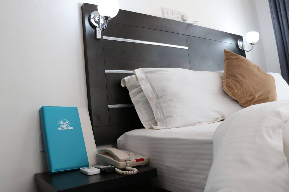 Uma cama bem arrumada com almofadas brancas e castanhas, uma mesa de cabeceira com telefone e controle remoto, e uma pasta azul no criado-mudo.