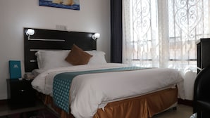 A neatly made bed with a headboard, a bedside table with a lamp, and a painting on the wall. - Fantasy Village Hotel (Dodoma)
