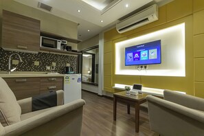 A modern hotel room with a kitchenette, a flat-screen TV mounted on the wall, and a seating area with a small table.