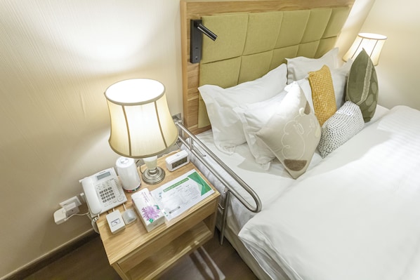 A hotel room with a bed, bedside table with a lamp, a phone, and a small bedside table with a tissue box and a brochure.