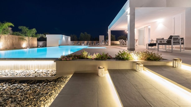 Un área moderna de piscina al aire libre con una piscina azul cristalina, rodeada por un solárium de piedra y un muro bajo con plantas. Hay sillones de descanso y una mesa bajo una zona cubierta.