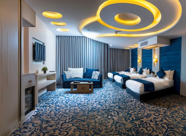Una habitación de hotel moderna con una cama grande, un sofá, una mesita y un televisor. La habitación tiene una alfombra estampada y un techo con iluminación circular.