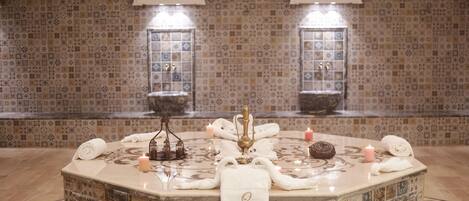 Un espacio de spa con pared de azulejos, una fuente central y toallas sobre una encimera de mármol.