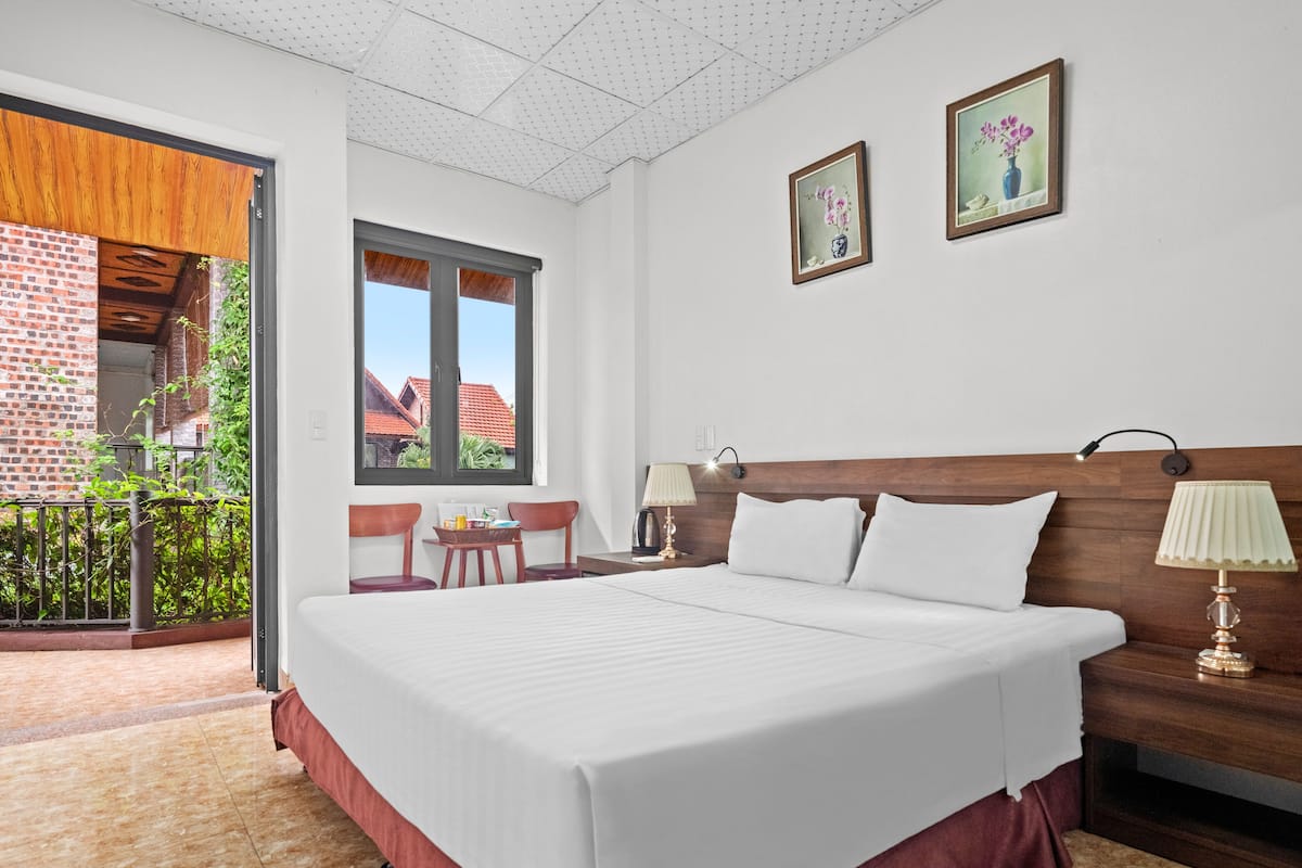 A hotel room with a large bed, two bedside tables with lamps, a window with a view of buildings, and a balcony with chairs and a table.