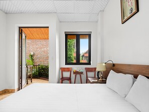 A hotel room with a bed, a desk with a lamp, a chair, and a window with a view of a brick building. - Tam Coc Haven Boutique Ninh Binh (Nam Hoa Lu)