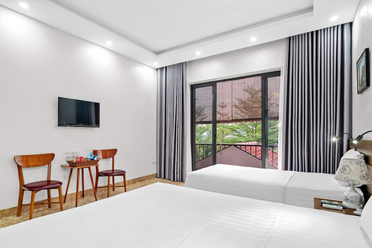 A hotel room with a large bed, a TV mounted on the wall, a small table with chairs, and a window with curtains.