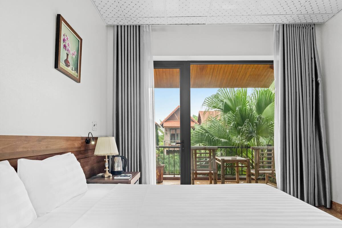 A hotel room with a large bed, a nightstand with a lamp, a framed picture on the wall, and a balcony with a table and chairs.