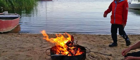 Escena de playa con una fogata, botes y una persona con una chaqueta roja.