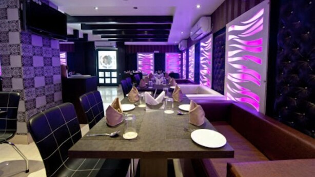 Interior de restaurante com padrão quadriculado na parede, iluminação ambiente roxa e área de estar com mesas preparadas para refeições.