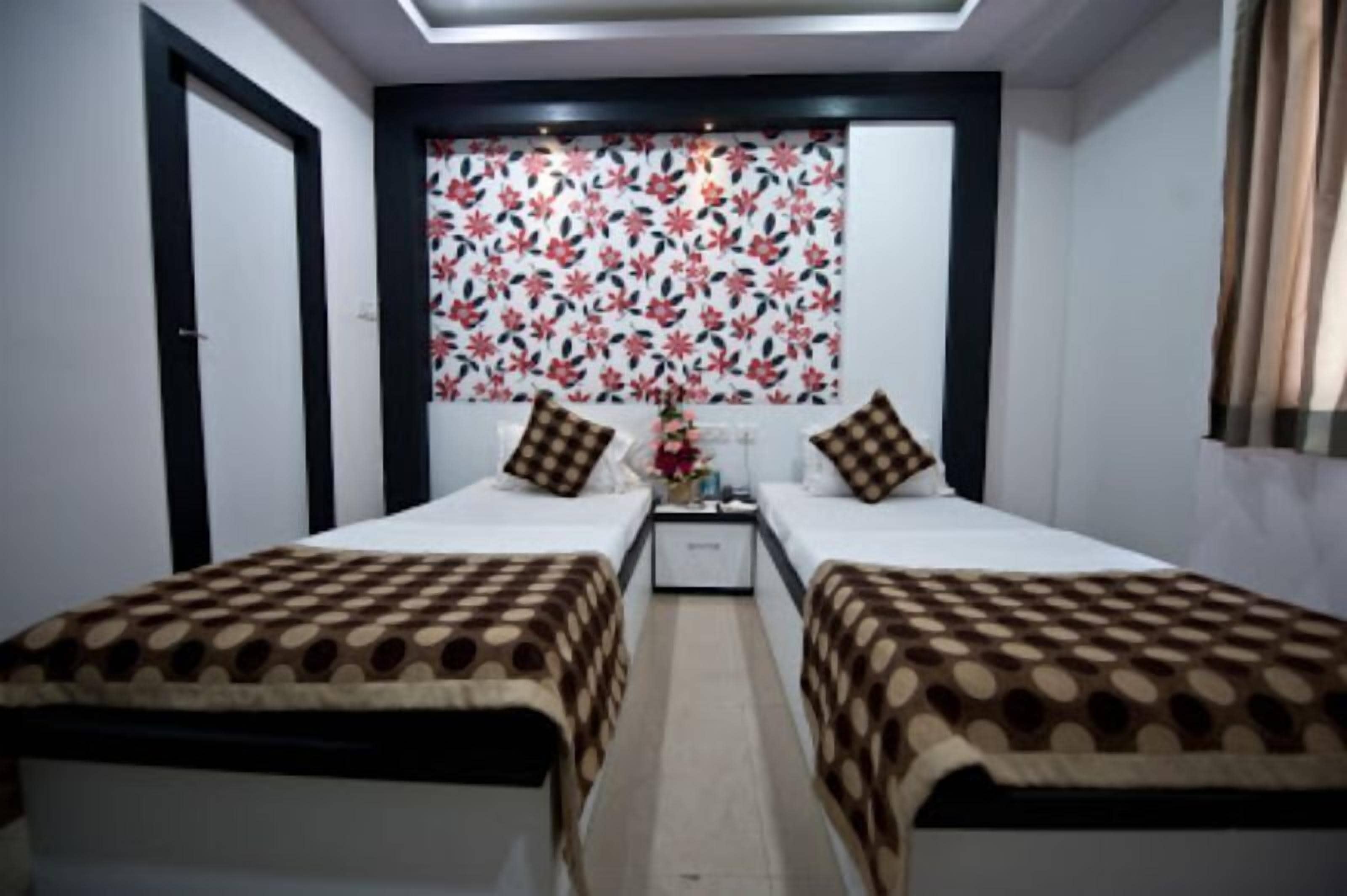A hotel room with two single beds, a nightstand, a small table with a vase, and a floral-patterned curtain.