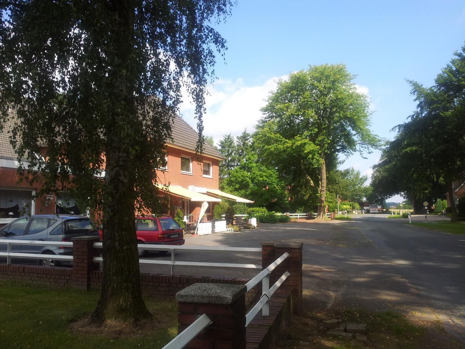 A residential street with parked cars, a brick house, and a large tree.